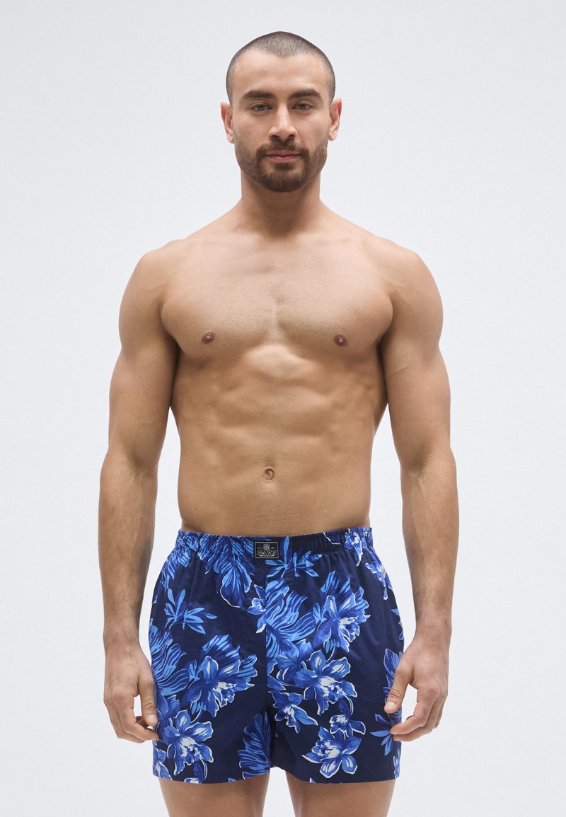 Blue floral patterned swim shorts made of lightweight fabric, featuring an elastic waistband and loose fit. Navy background with bright blue flowers.