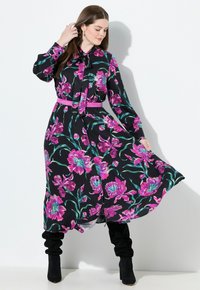 Kvinde iført en sort langærmet blomstret kjole med pink og grønne blomster, et pink bælte og sorte slouchy støvler, som poserer mod en hvid væg.