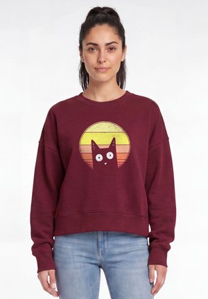 Frau mit dunklen, zusammengebundenen Haaren, trägt ein bordeauxfarbenes Sweatshirt mit einem schwarzen Katzenmotiv und Sonnenuntergangsstreifen, kombiniert mit hellblauen Jeans.
