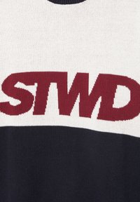 Zbliżenie tkaniny z wyraźnymi bordowymi literami „STWD” na białym górnym i czarnym dolnym dzianym tle.