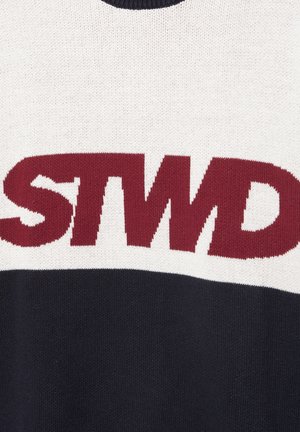 Közelkép egy szövetből, amelyen vastag mályvaszínű "STWD" betűk láthatók fehér felső és fekete alsó kötött háttéren.