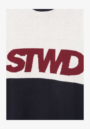 Közelkép egy szövetből, amelyen vastag mályvaszínű "STWD" betűk láthatók fehér felső és fekete alsó kötött háttéren.