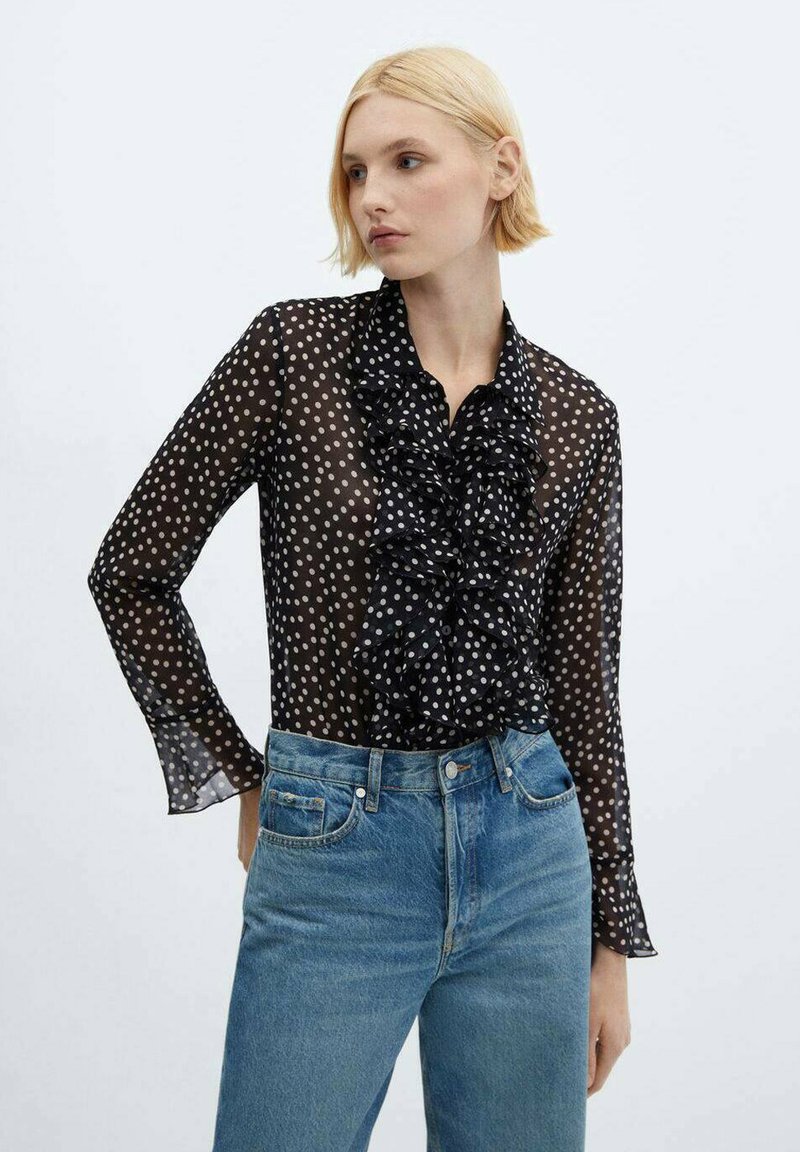 Frau trägt eine durchsichtige schwarze Bluse mit langen Ärmeln, weißen Punkten und Rüschendetails, kombiniert mit hochgeschnittenen blauen Jeans.