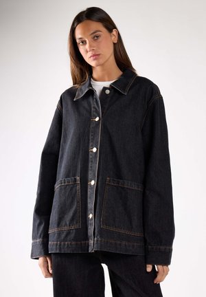 Frau trägt eine übergroße schwarze Jeansjacke mit Fronttaschen und weißen Knöpfen über einem weißen Hemd, stehend vor einem einfarbigen Hintergrund.