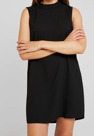 Femme portant une robe noire sans manches à col montant, bras croisés sur la poitrine, debout devant un fond gris uni.