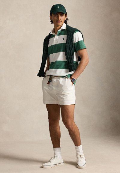 Jeune homme portant un polo rayé vert et blanc, un short blanc, des baskets blanches, une casquette verte et un pull foncé posé sur les épaules.