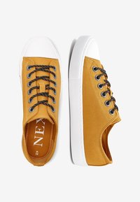 Žluté plátěné tenisky s texturovaným povrchem, bílé gumové podešve a černými tkaničkami. Mají zesílené očka a logo na stélce.