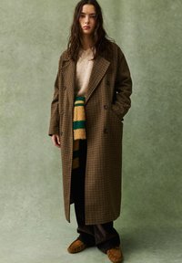 Junge Frau, die einen langen braunen karierte Mantel, einen beigen Pullover, einen gestreiften Schal, dunkle Hosen und braune Mokassins trägt, vor einem grünen Hintergrund.