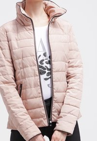 Kvinna klädd i en ljusrosa vadderad jacka med hög krage över en vit skjorta med svart leopardmönster och bokstavsdesign.