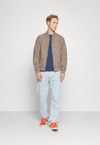Casaco bomber de camurça castanho, punhos ajustados, camisa azul-marinho, jeans azul-claro e ténis vermelhos. Design simples com detalhes em zíper.