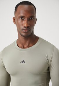 Long-sleeve athletic shirt in light olive green, made of stretch fabric. Features a round neckline and a black Adidas logo on the chest.