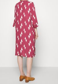 Kinono-stil klänning i burgundy tyg med vita triangulära mönster. Trekvartslånga ärmar och sidoklyftor. Kombinerad med vävda bruna skor.
