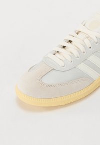Närbild av en beige och grå sneaker-tå med krämfärgade snören och gummisula, med tyg- och mockatexturer.