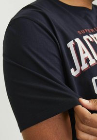 Mørkeblå bomulds T-shirt med korte ærmer, prydet med fed hvid og maroon trykt tekst "JACKSON" og "SUPERIOR" på brystet og skulderen.