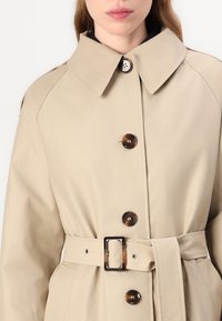 Béžový trenčkot s velkým límcem, s hnědými knoflíky a odpovídajícím páskem, vyrobený z hladkého materiálu.
