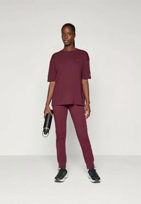 Burgunderfarget treningsantrekk bestående av en løs t-skjorte og tettsittende bukser, laget av jersey-stoff. Har en subtil logo og svarte joggesko.