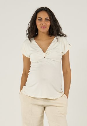 Frau mit langen dunklen Haaren, trägt ein cremefarbenes Kurzarmtop und hellbeige Hosen, steht mit den Händen in den Taschen vor einem einfarbigen Hintergrund.