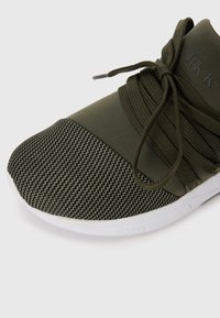 Chaussure de sport vert olive avec un bout en mesh texturé et un dessus lisse. Dotée de lacets élastiques et d'une semelle en caoutchouc blanche.