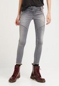 Jeans skinny grises con parches acolchados en las rodillas, cierre de doble botón y un ligero desvanecimiento, combinados con botas burdeos de cordones.