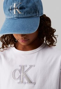 Cappello in denim di colore azzurro chiaro con logo ricamato "Calvin Klein". T-shirt bianca con grande logo "CK" ricamato in argento.