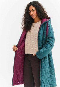 Dlouhý prošívaný kabát v barvě teal s vínově červenou vnitřní podšívkou. Kabát má kapuci, zapínání na zip a prošívání v diamantovém vzoru. Pod ním je béžový pletený svetr.