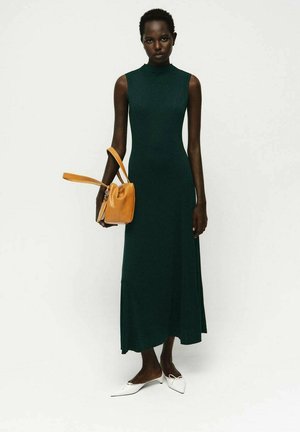 Abito lungo verde senza maniche con scollatura alta, abbinato a una piccola borsa gialla e scarpe bianche con punta appuntita. Tessuto liscio, silhouette aderente.