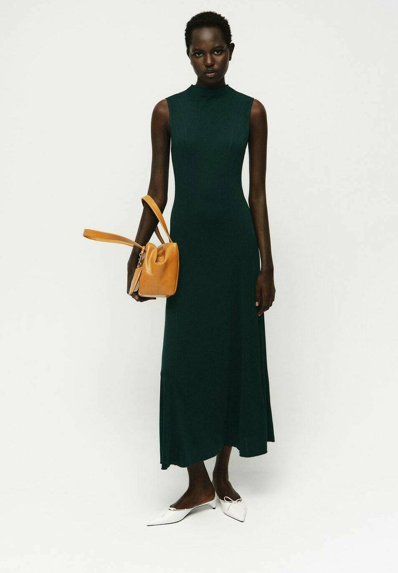 Abito lungo verde senza maniche con scollatura alta, abbinato a una piccola borsa gialla e scarpe bianche con punta appuntita. Tessuto liscio, silhouette aderente.