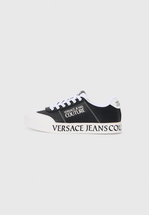 Svart sneakers med teksturert overflate, hvite snørebånd, og en fremtredende hvit såle. Merking "VERSACE JEANS COUTURE" på siden og hælen.