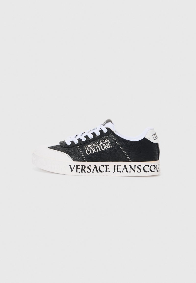 Svart sneakers med teksturert overflate, hvite snørebånd, og en fremtredende hvit såle. Merking "VERSACE JEANS COUTURE" på siden og hælen.