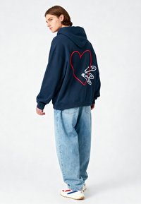 Felpa oversized blu navy con un cuore rosso e spille di sicurezza bianche cucite sul retro, abbinata a jeans baggy blu chiaro e sneakers.