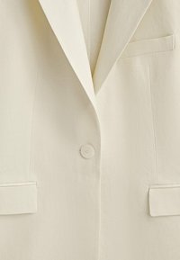 Cremefarbener Blazer mit Reverskragen, Ein-Knopf-Verschluss und zwei Vorderseiten-Taschen. Glatter Stoff mit weicher Textur.