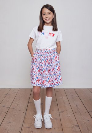 Ragazza sorridente con capelli castani lisci, mani in tasca, indossa una maglietta bianca ESCADA, gonna multicolore ESCADA, calzini bianchi e scarpe da ginnastica bianche.