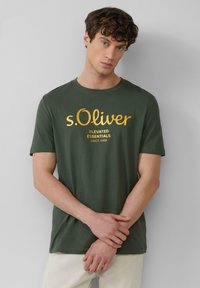 Junger Mann trägt ein dunkelgrünes s.Oliver-T-Shirt mit gelber Schrift und steht vor einem schlichten hellgrauen Hintergrund, die Hände locker gefaltet.