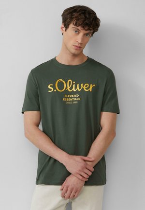 Een jonge man in een donkergroene s.Oliver t-shirt met gele tekst, staande tegen een effen lichtgrijze achtergrond, met de handen losjes gekruist.