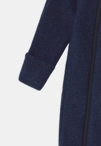 Macacão de polar azul marinho com uma superfície texturizada; apresenta um fecho éclair na parte da frente e punhos canelados nas mangas.