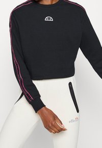 Korte zwarte sweatshirt met roze zijstrepen en een wit logo op de borst, gecombineerd met lichte joggingsbroek met ritszakken.