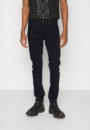 Mężczyzna w czarnych dopasowanych dżinsach, czarnej koszulce z nadrukiem i czarnych skórzanych butach na sznurowadła, stojący na jednolitym tle.