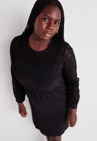 Robe noire avec un corsage et des manches en dentelle, un col rond et une taille cintrée. Le tissu a une texture lisse avec des motifs en dentelle élaborés.