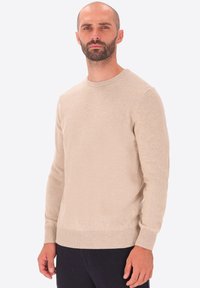 Uomo con barba e capelli corti, che indossa un maglione beige a girocollo e pantaloni neri, in piedi davanti a uno sfondo bianco semplice.