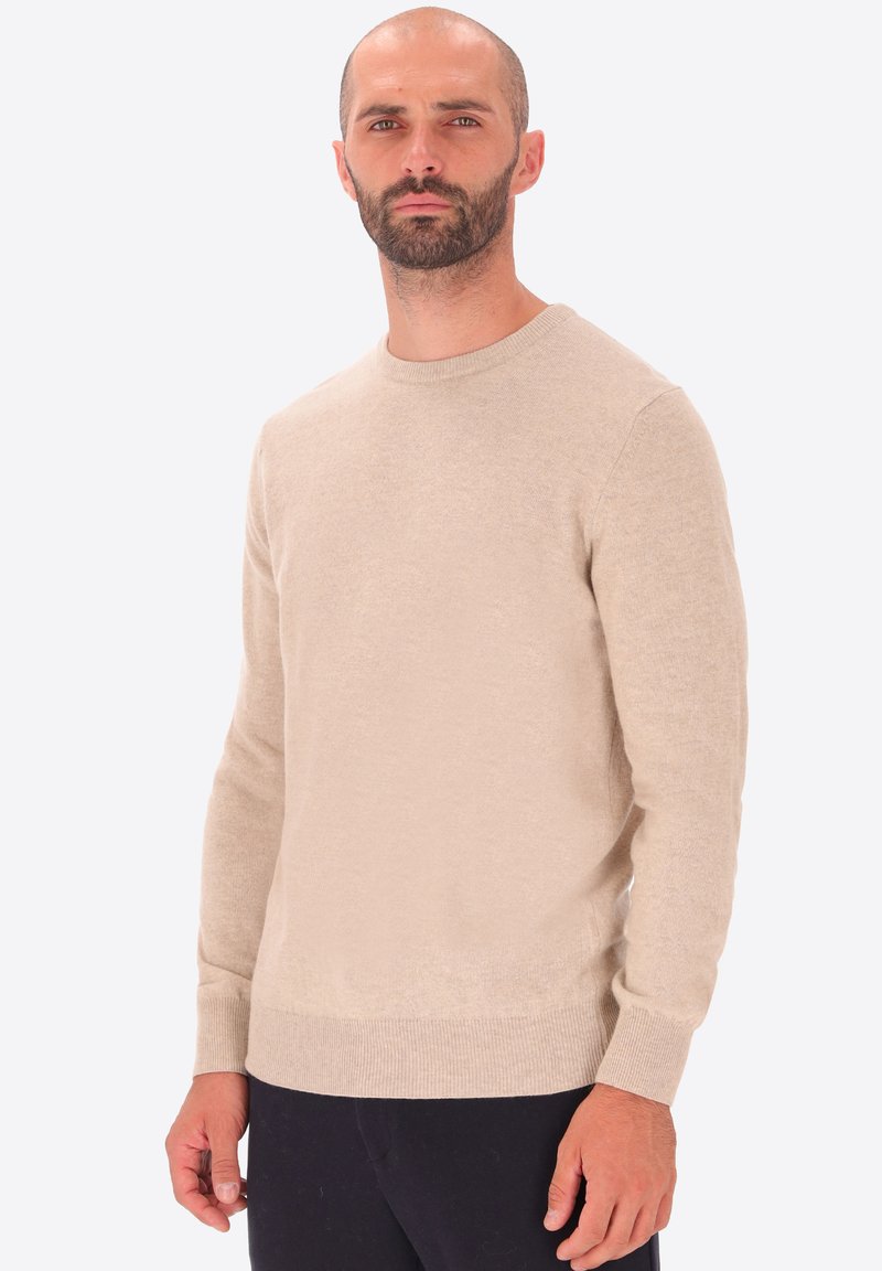 Uomo con barba e capelli corti, che indossa un maglione beige a girocollo e pantaloni neri, in piedi davanti a uno sfondo bianco semplice.