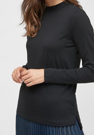 Femme portant un haut noir à manches longues avec col montant factice et une jupe bleue plissée, debout les mains légèrement croisées devant elle.