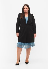 Zwarte blazer met een lange, getailleerde silhoutte, gecombineerd met een lichtblauw kantendraag met een golvende zoom. Zwarte hoge hakken maken de look compleet.