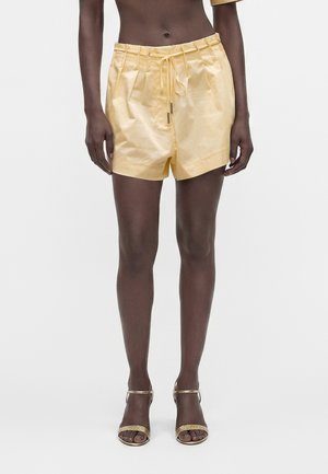 Personne portant un short plissé jaune pâle avec cordon de serrage et talons ouverts dorés, debout devant un fond clair uni.