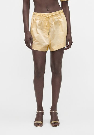 Personne portant un short plissé jaune pâle avec cordon de serrage et talons ouverts dorés, debout devant un fond clair uni.