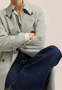 Jasnoszary trencz o podwójnym zapięciu, z czarnymi guzikami, paskiem w talii i teksturowanymi rękawami, zestawiony z ciemnoniebieskimi dżinsami.