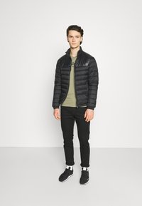 Svart pufferjakke med quiltet design og ribbede mansjetter, iført over en grønn t-skjorte, kombinert med svarte bukser og svarte sneakers.