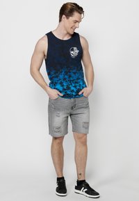 Modra gradient majica brez rokavov z abstract vzorcem in grafičnim tiskom na žepu, v kombinaciji s sivimi uničenimi jeans kratkimi hlačami in črnimi supergami.