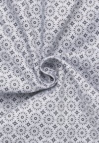 Weisser Stoff mit einer wirbelnden Falte, die ein sich wiederholendes Muster aus kleinen schwarzen floralen und geometrischen Formen zeigt.