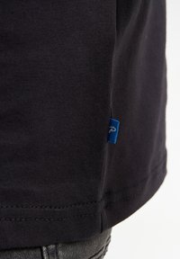 Camiseta de algodón negra con una textura acanalada, que presenta una pequeña etiqueta azul en la costura lateral, destacando un acabado limpio con costuras dobles.