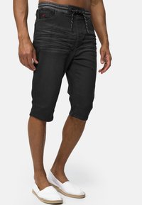 Shorts de mezclilla negra con una cintura elástica gris, que presentan pliegues laterales y un cordón, llevados con zapatos blancos tipo slip-on.